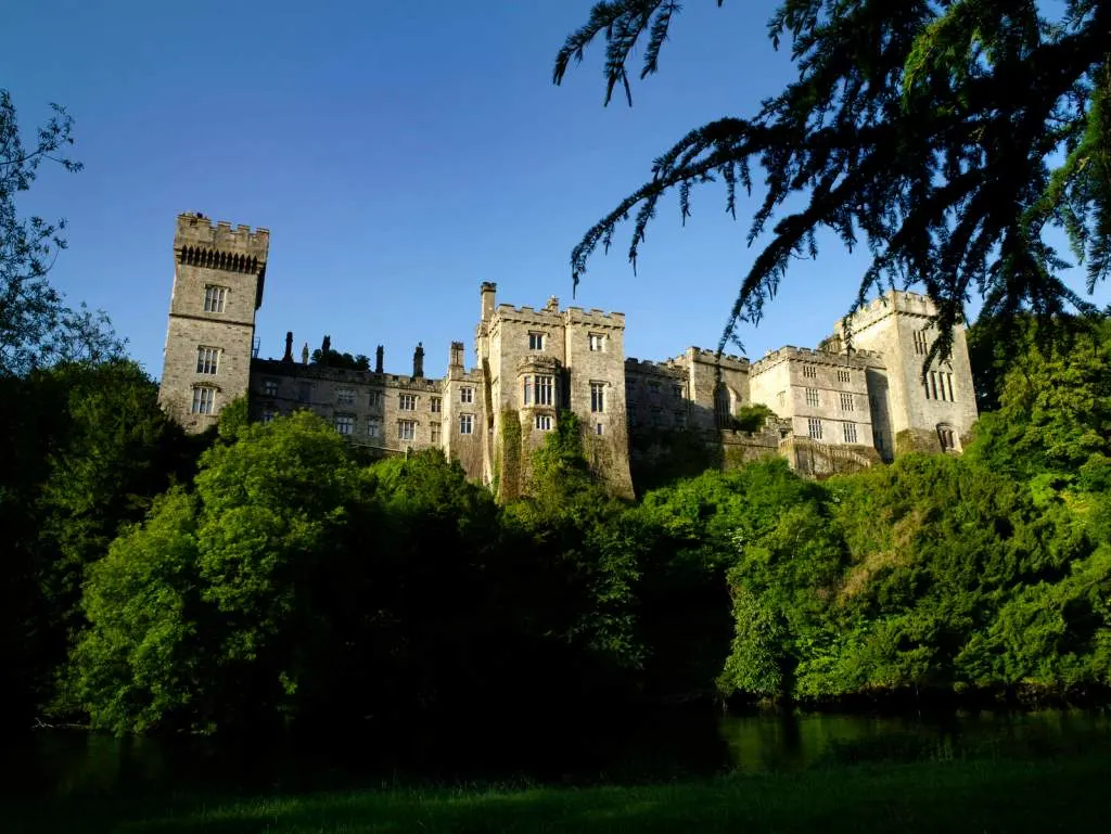 Medieval castle with multiple stone towers surrounded by dense green trees beside river