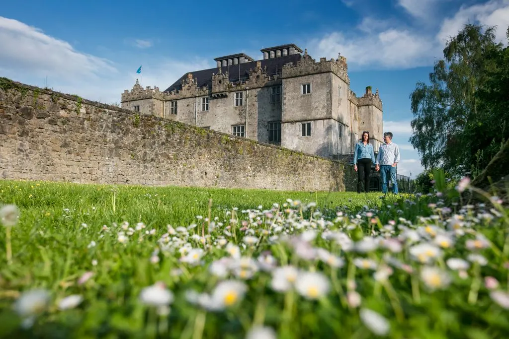 Two visitors walk on grassy grounds near a stone castle with manicured lawns and white flowers in foreground