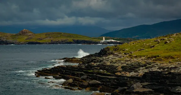 Rocky coastline with green hills, lighthouse, and stormy sky over Atlantic waters