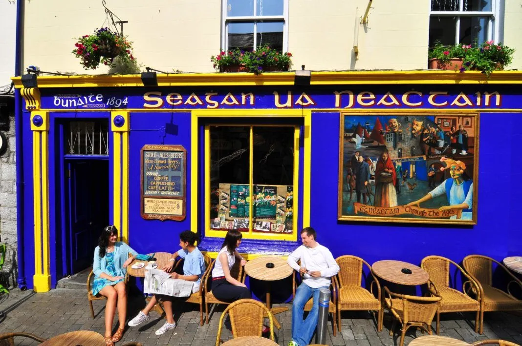 Visitors seated outside Tigh Neachtain's blue and yellow pub front in Galway, with hanging flower baskets