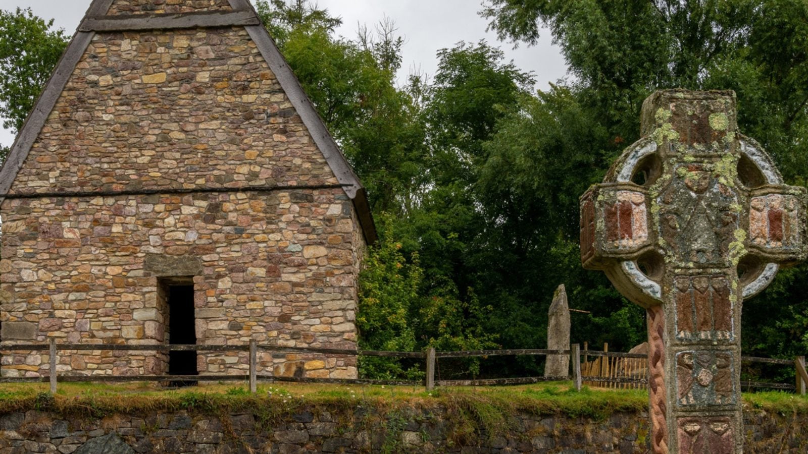 Ancient Celtic cross with spiral carvings beside a stone chapel at the Irish National Heritage Park, representing the era of Brehon Law in Ireland