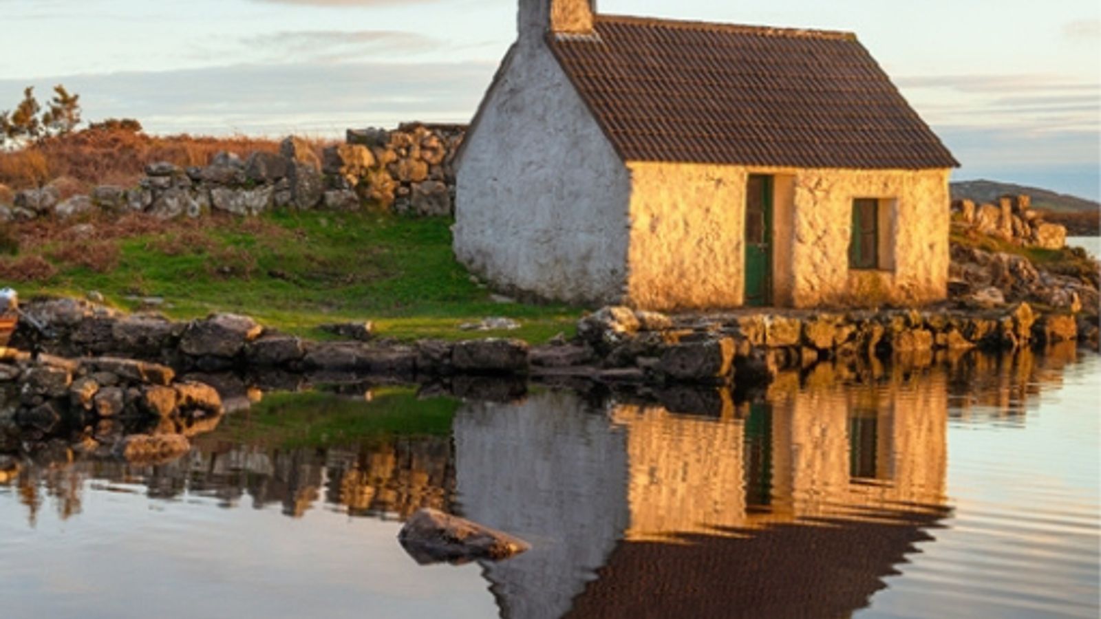 A traditional Irish stone cottage reflected in still water at golden sunset, Connemara, Ireland