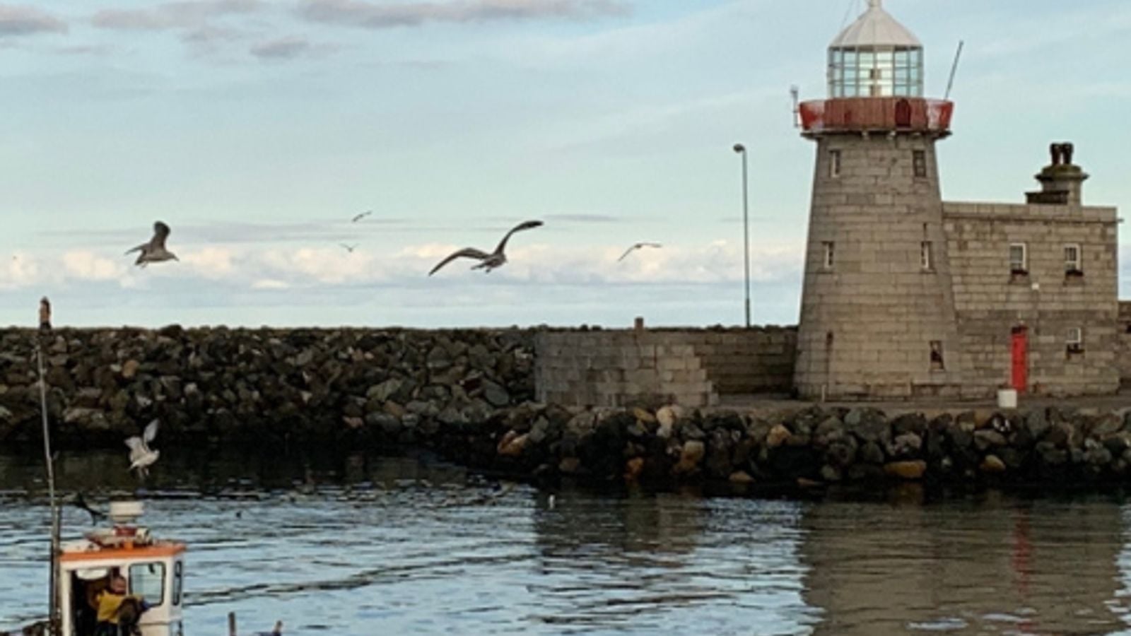 A small fishing boat returning to Howth Harbour past the lighthouse at dusk, seagulls overhead