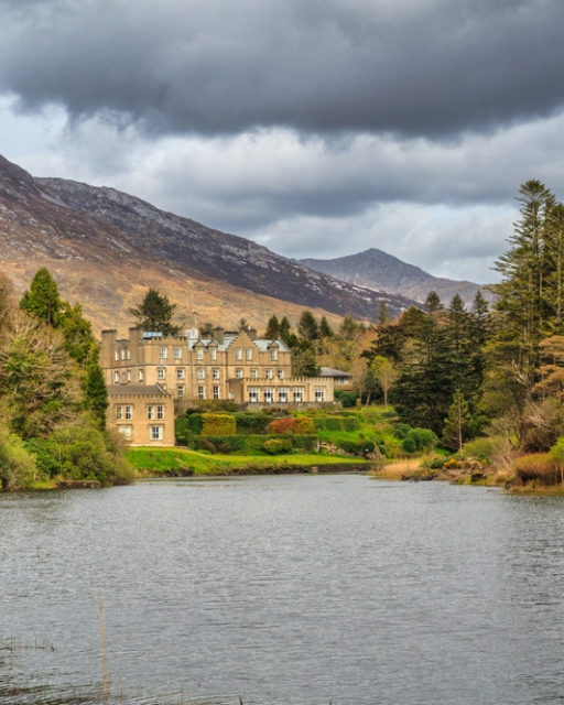 Historic stone castle beside a lake with mountains and evergreen trees under cloudy sky