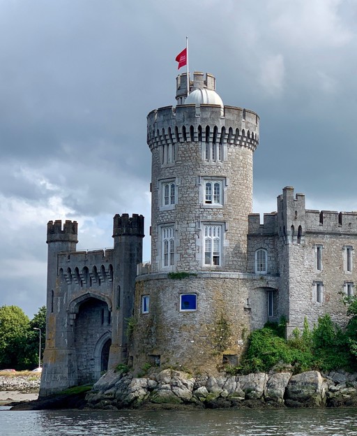 Round stone castle tower with crenellations beside water, Irish flag flying