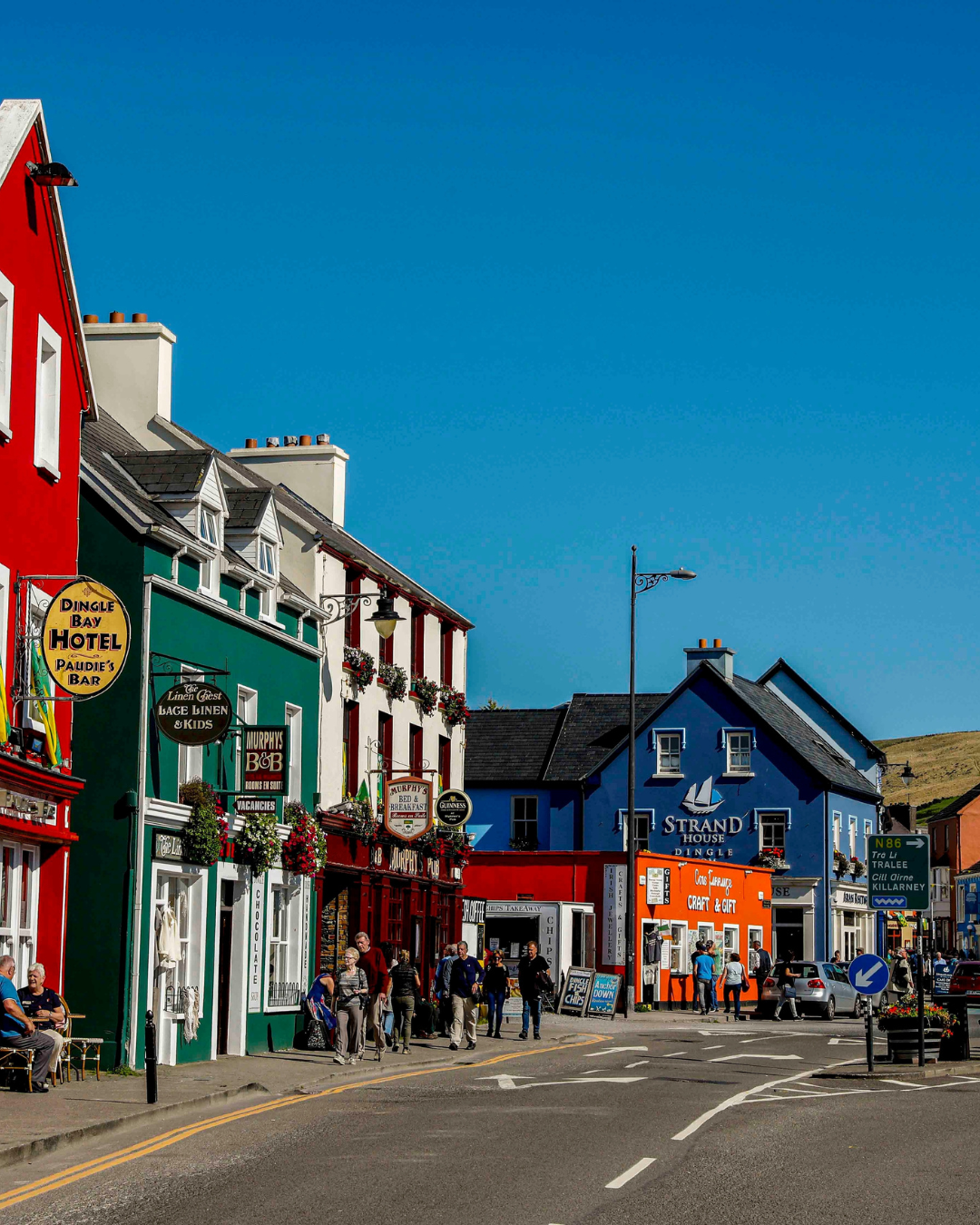 Colourful shopfronts lining a sunny street in Dingle, County Kerry, Ireland, with bright red, green, and blue painted facades