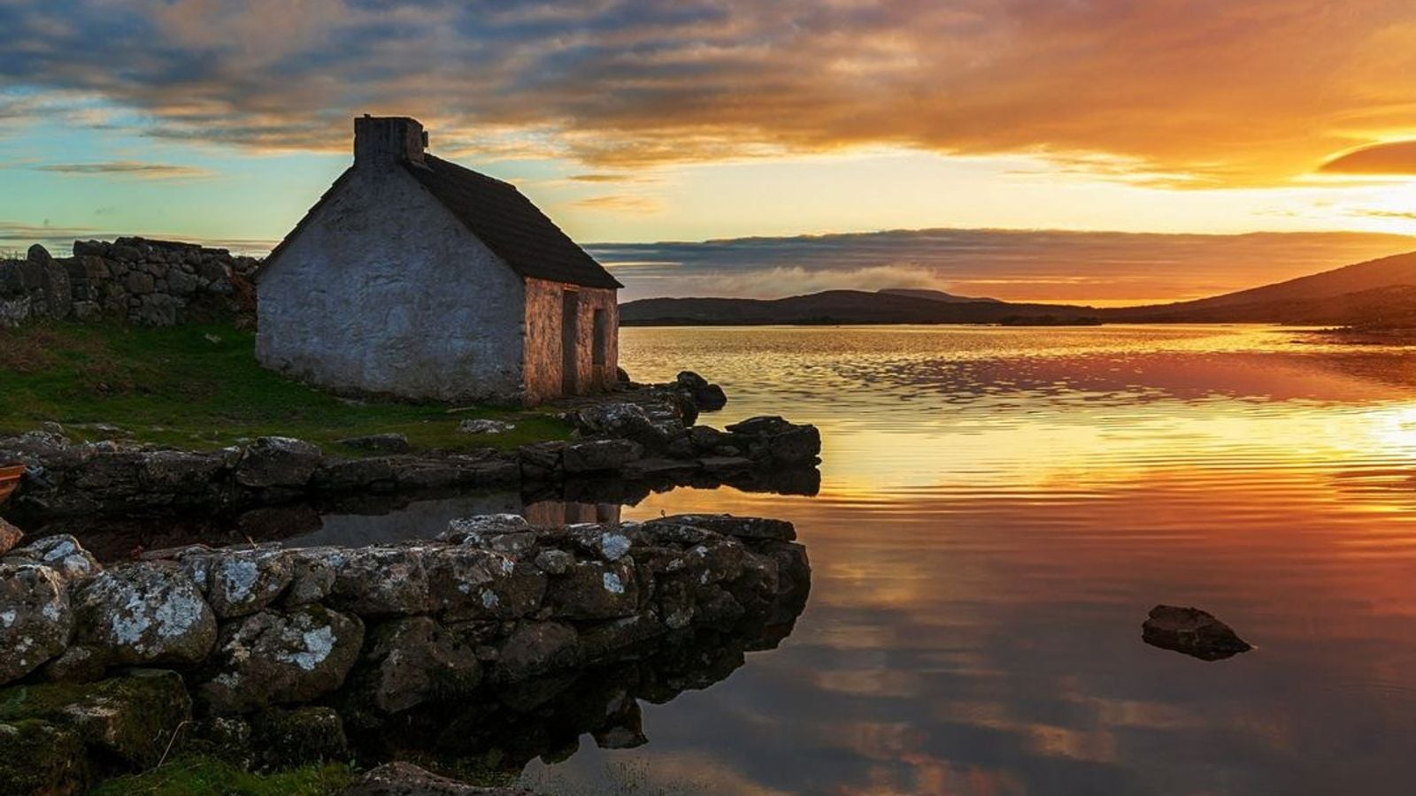 Traditional Irish stone cottage at sunset beside a calm lake in Connemara, County Galway, Ireland