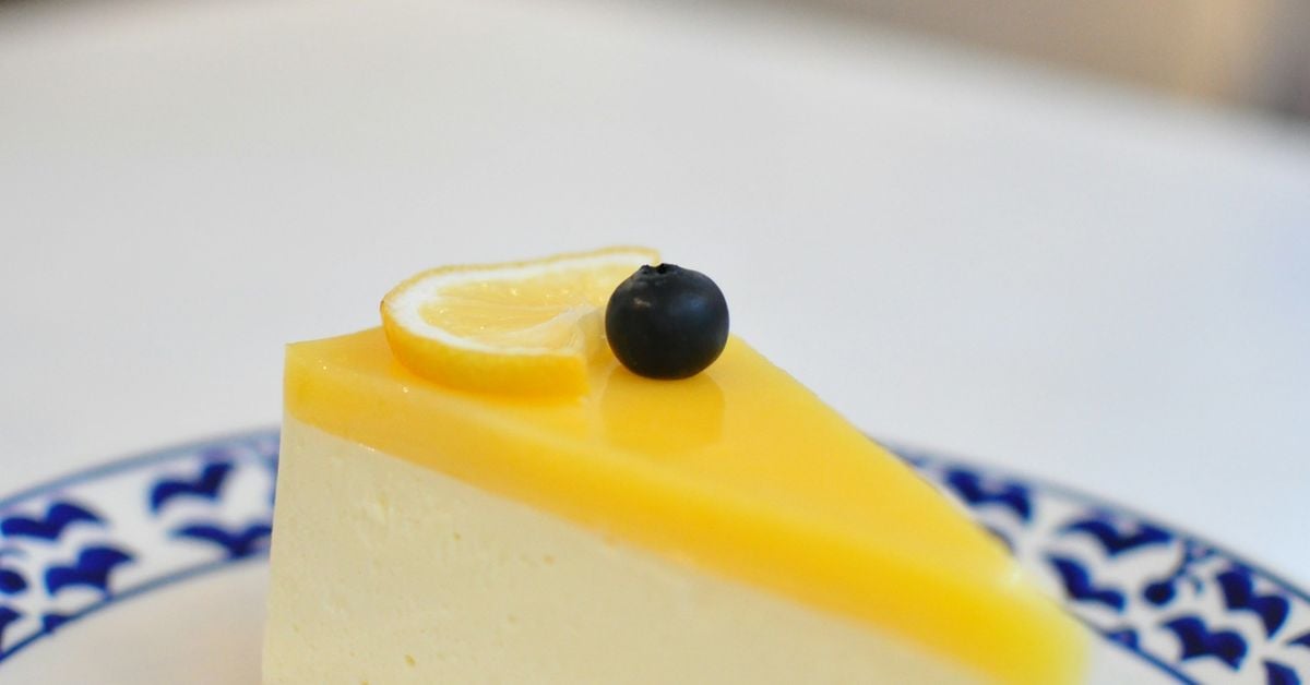 A slice of lemon cheesecake with creamy yellow filling on a digestive biscuit base, garnished with a lemon slice and blueberry