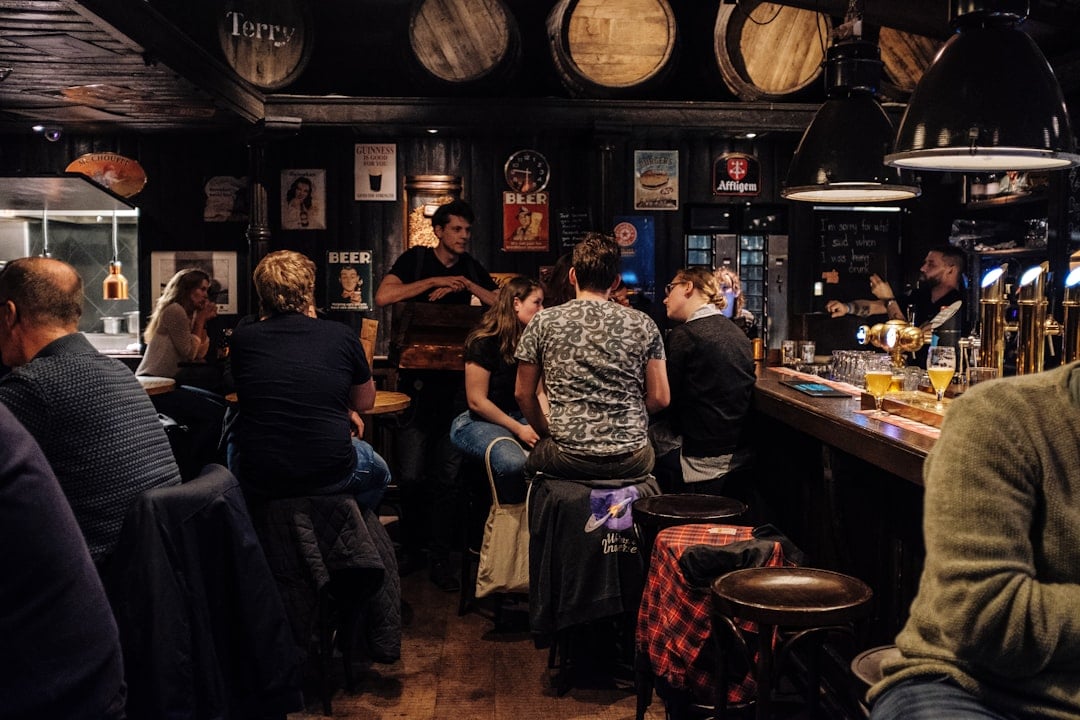People gathering at a dimly lit pub bar with wooden barrels overhead and vintage beer signage