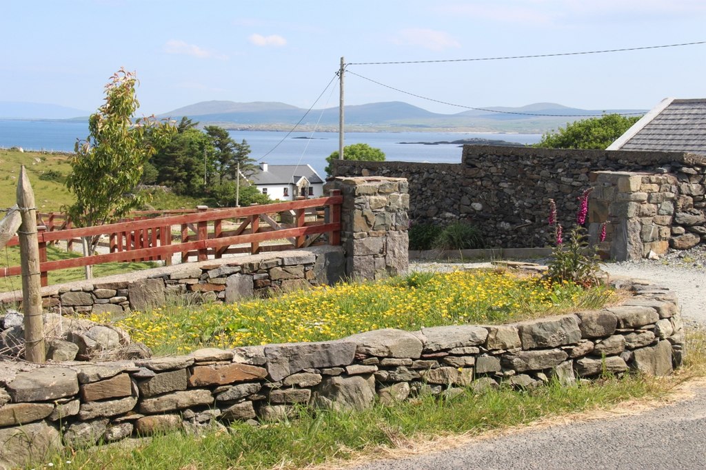 Traditional stone-walled cottage smallholding on the Connemara loop road in the west of Ireland, with a red timber gate, wildflowers, a lake, and mountains behind