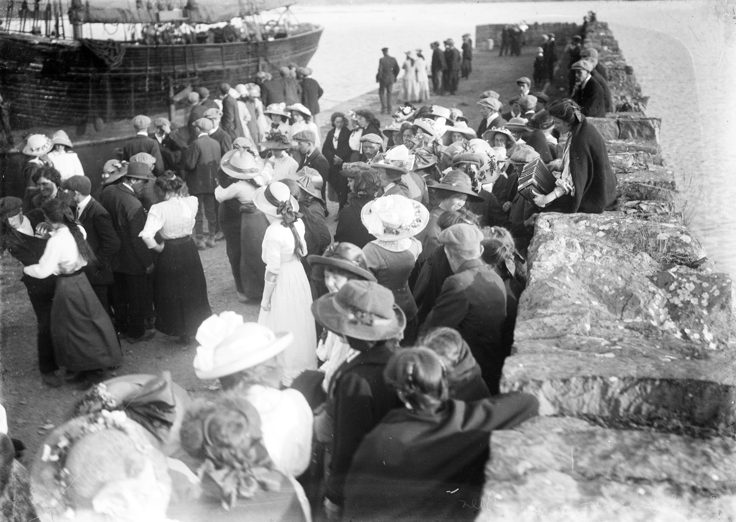 Vintage photo of a melodeon player on a stone wall and a crowd dancing outdoors during the Kilmakilloge Pattern in County Kerry, Ireland — the same outdoor music-and-dance tradition later practiced at rural crossroads