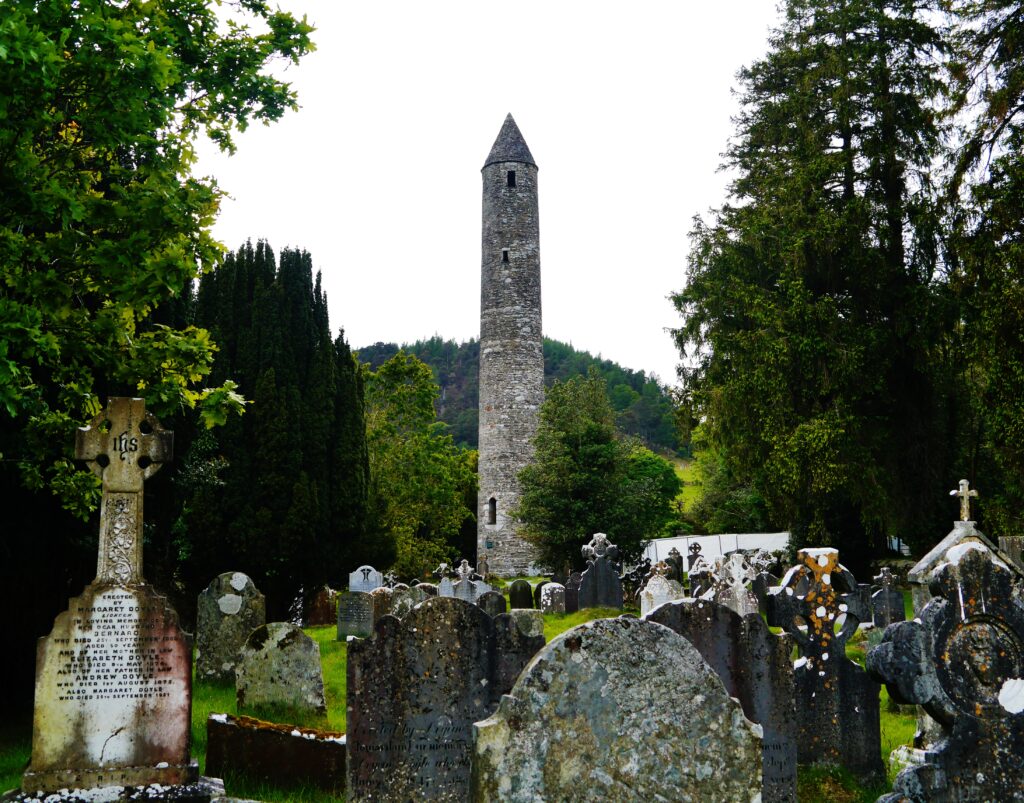 Celtic crosses and gravestones in Glendalough cemetery, County Wicklow, with the medieval round tower rising behind the trees