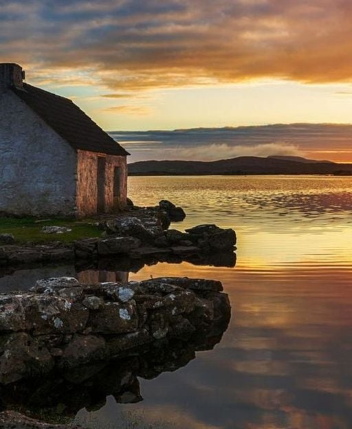 A stone cottage on a Connemara lake shore at sunset, golden light over the water and hills