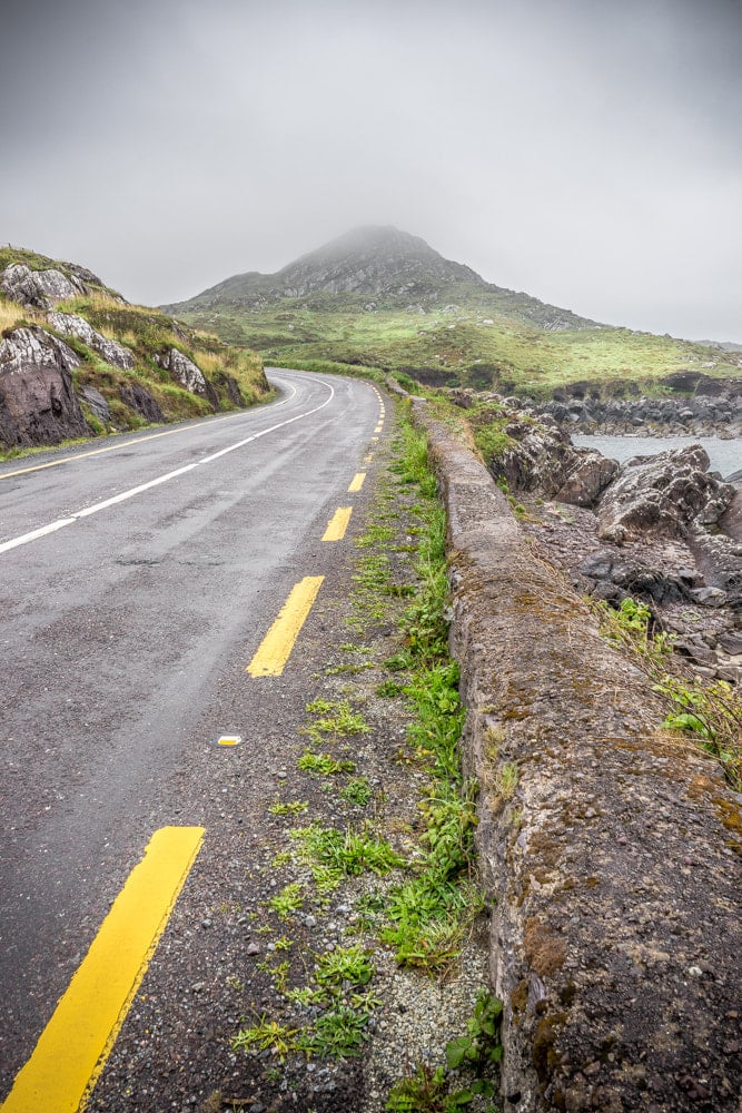 A coastal road winding along the Ring of Kerry with a fog-covered mountain rising ahead, illustrating how Ireland's coast and mountain landscapes meet
