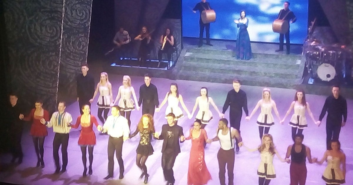 Riverdance ensemble dancers linked arm-in-arm on stage during a curtain call, with musicians and a vocalist performing on a platform behind them