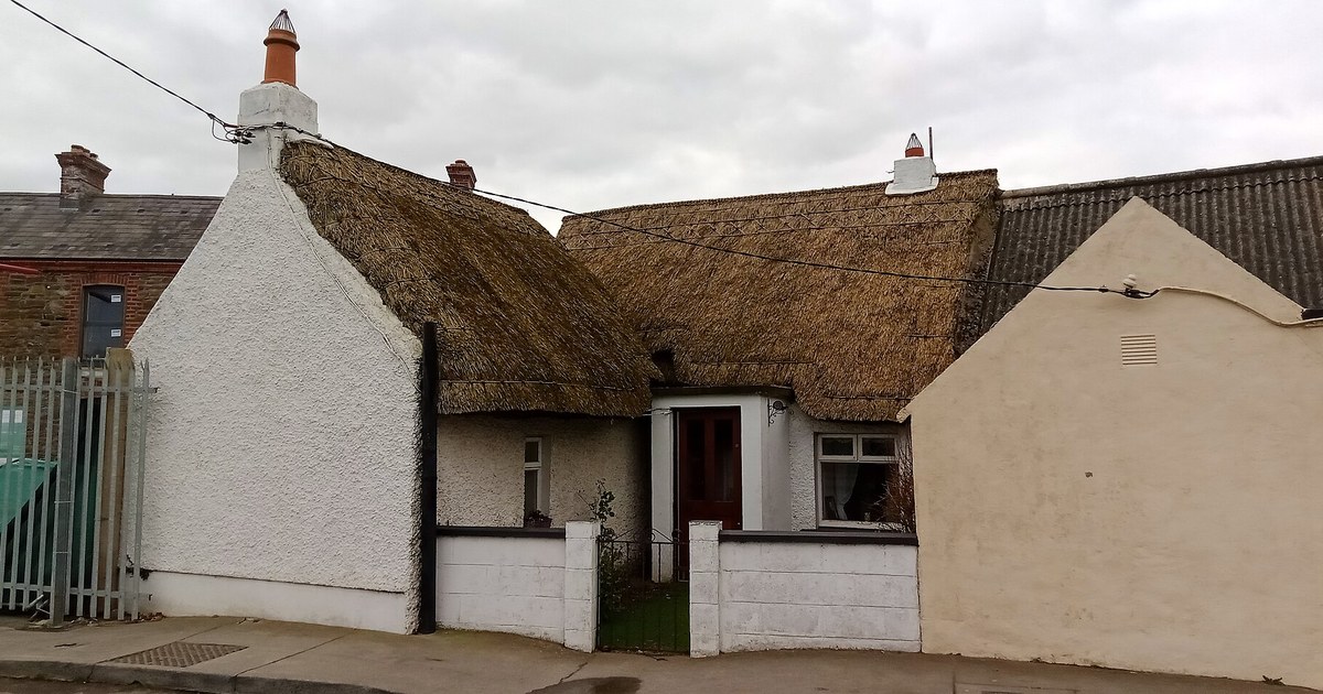 A whitewashed, thatched Irish cottage with two chimney pots and a low front wall, squeezed between taller modern buildings on a town street
