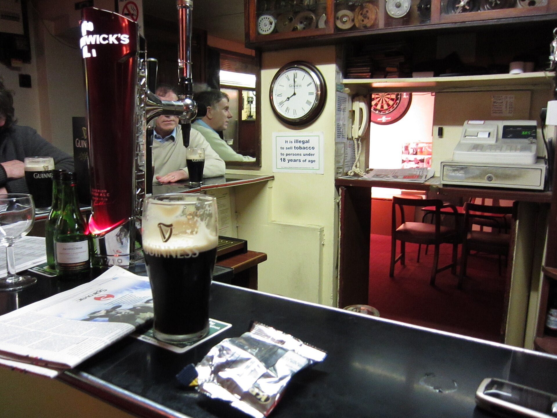 Interior of a traditional Irish pub, with a fresh pint of Guinness on the bar, a Guinness wall clock, and a dartboard visible through to the next room