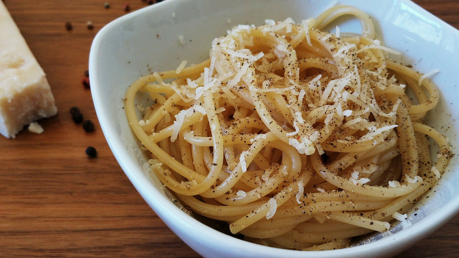Bowl of spaghetti cacio e pepe topped with grated pecorino romano and cracked black pepper, with a wedge of pecorino and peppercorns alongside
