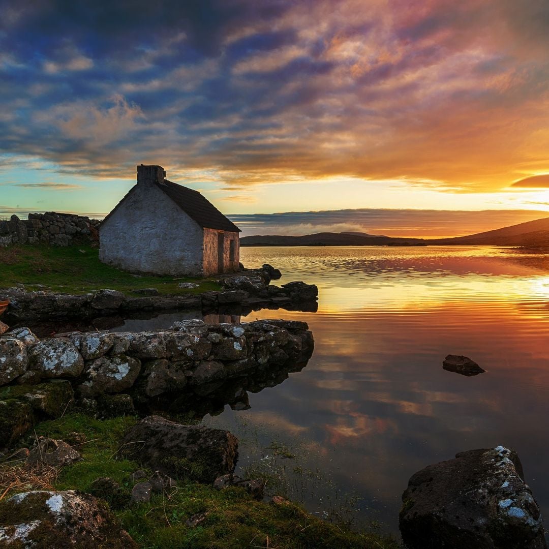 A traditional whitewashed stone cottage on the shore of a Connemara lough at sunset, with dramatic orange and blue clouds reflected in the still water