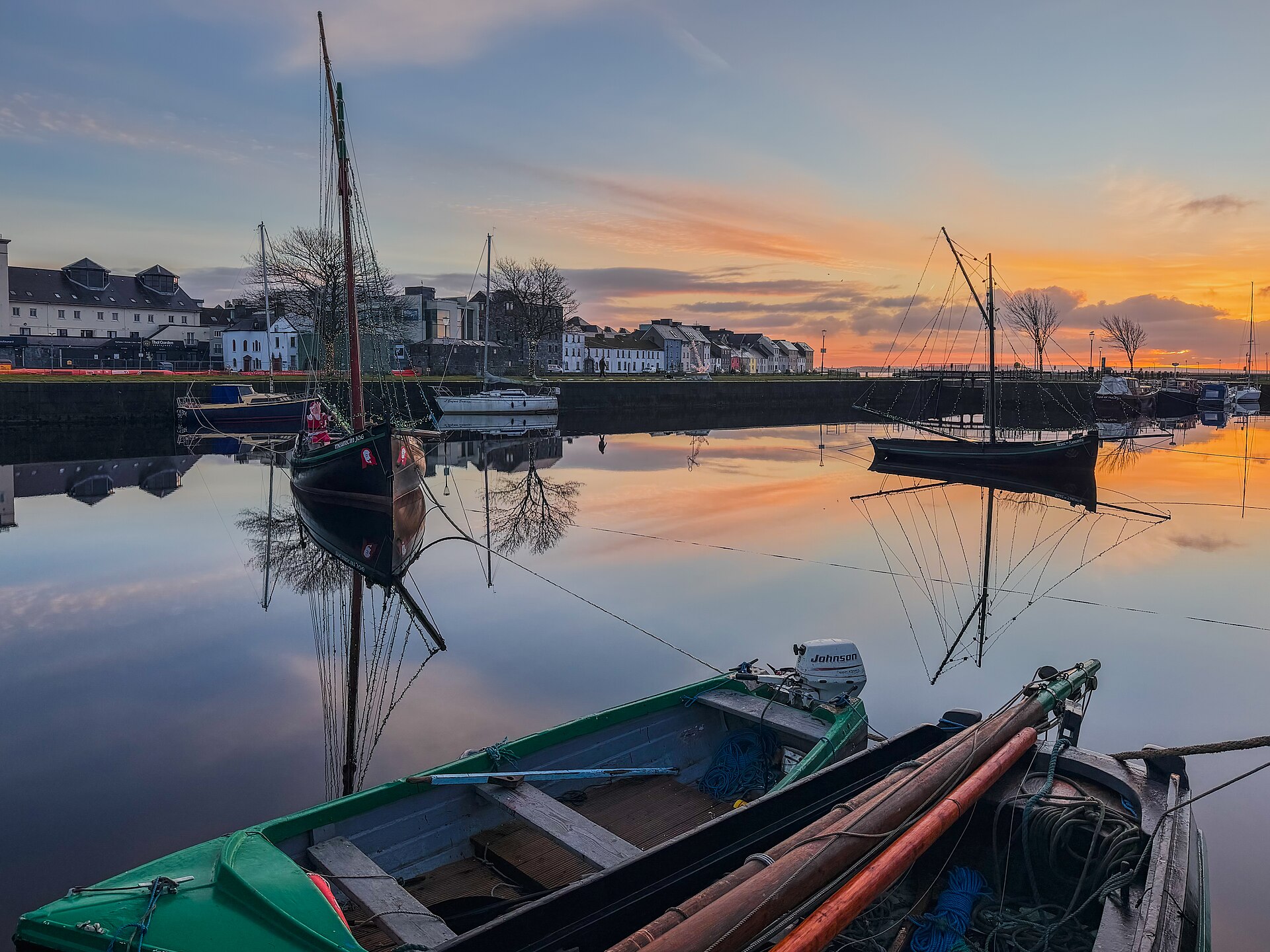 Galway hookers with dark hulls and tall masts moored in Galway harbour at sunrise, with the Long Walk houses behind