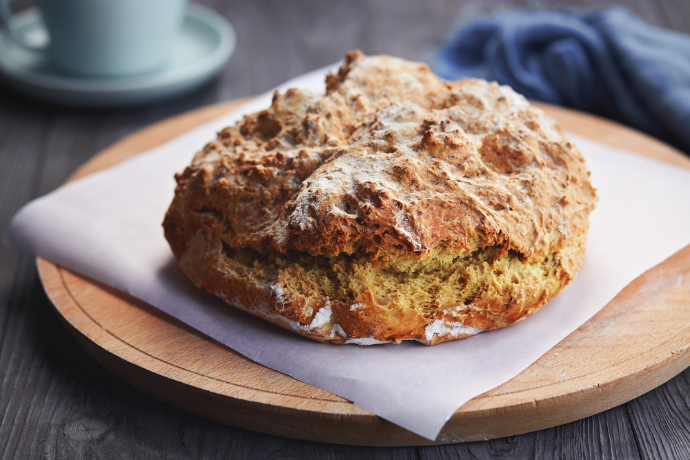 A round Irish brown soda bread loaf with a deeply browned, cracked crust, sliced open on a wooden board