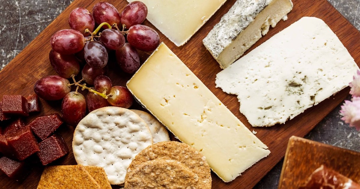 Wooden board with three farmhouse cheeses, red grapes, quince paste cubes and crackers, seen from above