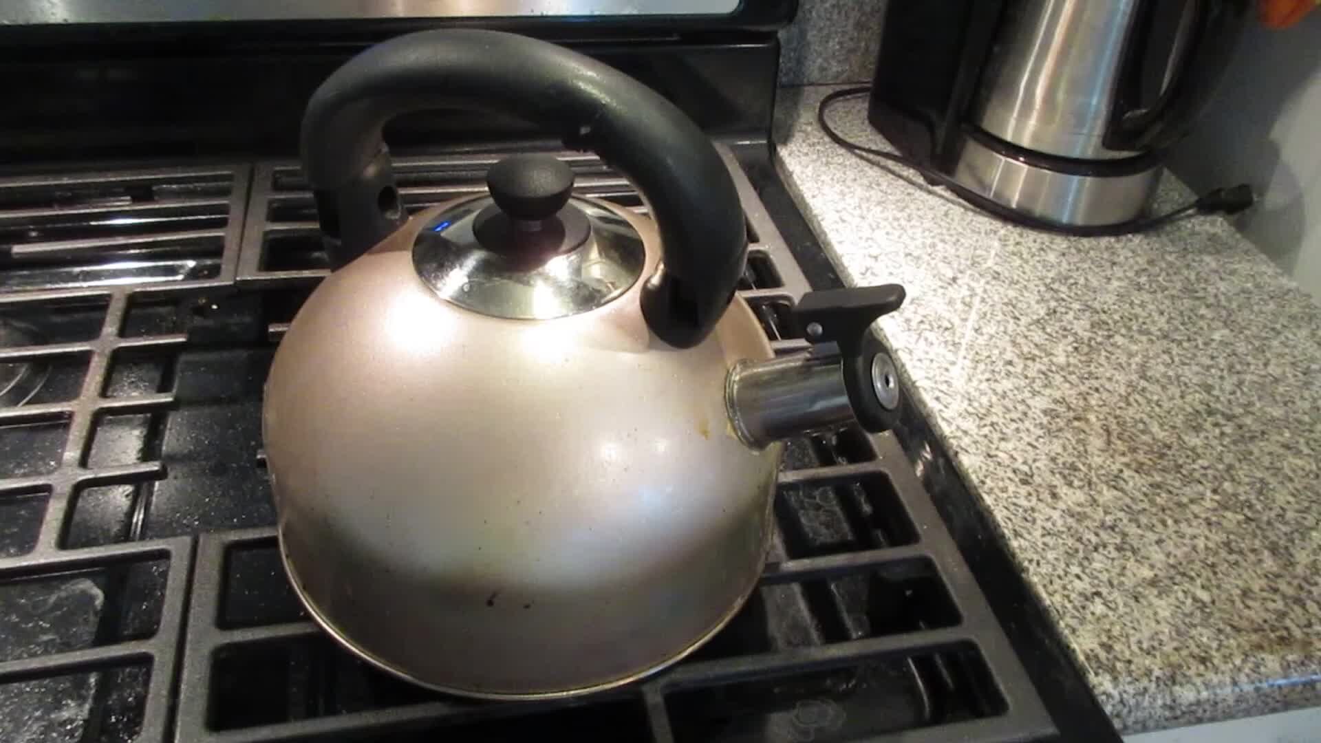 A stainless steel whistling kettle with a black handle sitting on a gas stove burner, the first thing put on in an Irish home during a crisis