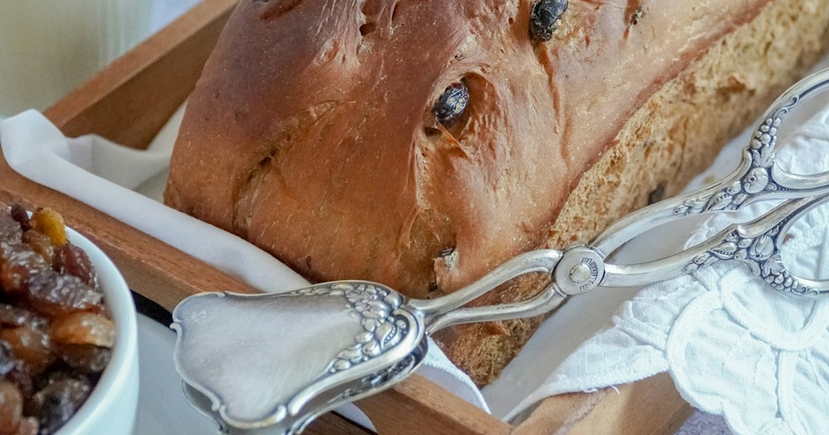 Freshly baked tea brack loaf studded with dried fruit, resting on a wooden board beside vintage silver pastry tongs and a bowl of soaked mixed fruit, wrapped in white linen