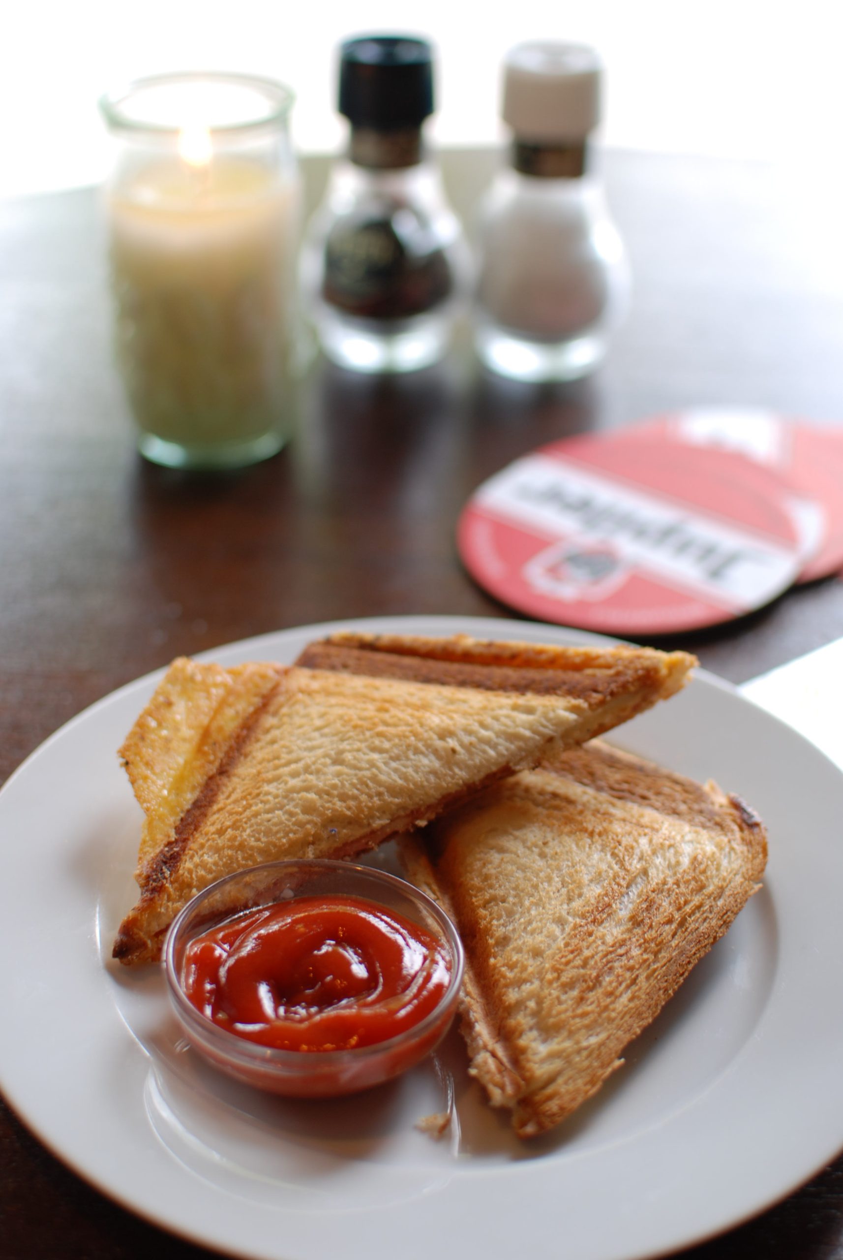 Golden-brown toasted ham and cheese sandwich cut into triangles on a plate with a small pot of ketchup, served on a caf&eacute; table