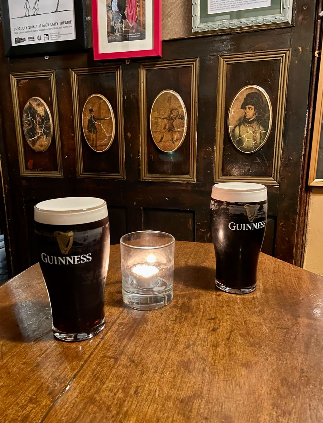 Two pints of Guinness and a lit tealight on a wooden table in a traditional Irish pub, with old framed pictures on the wood-panelled wall behind