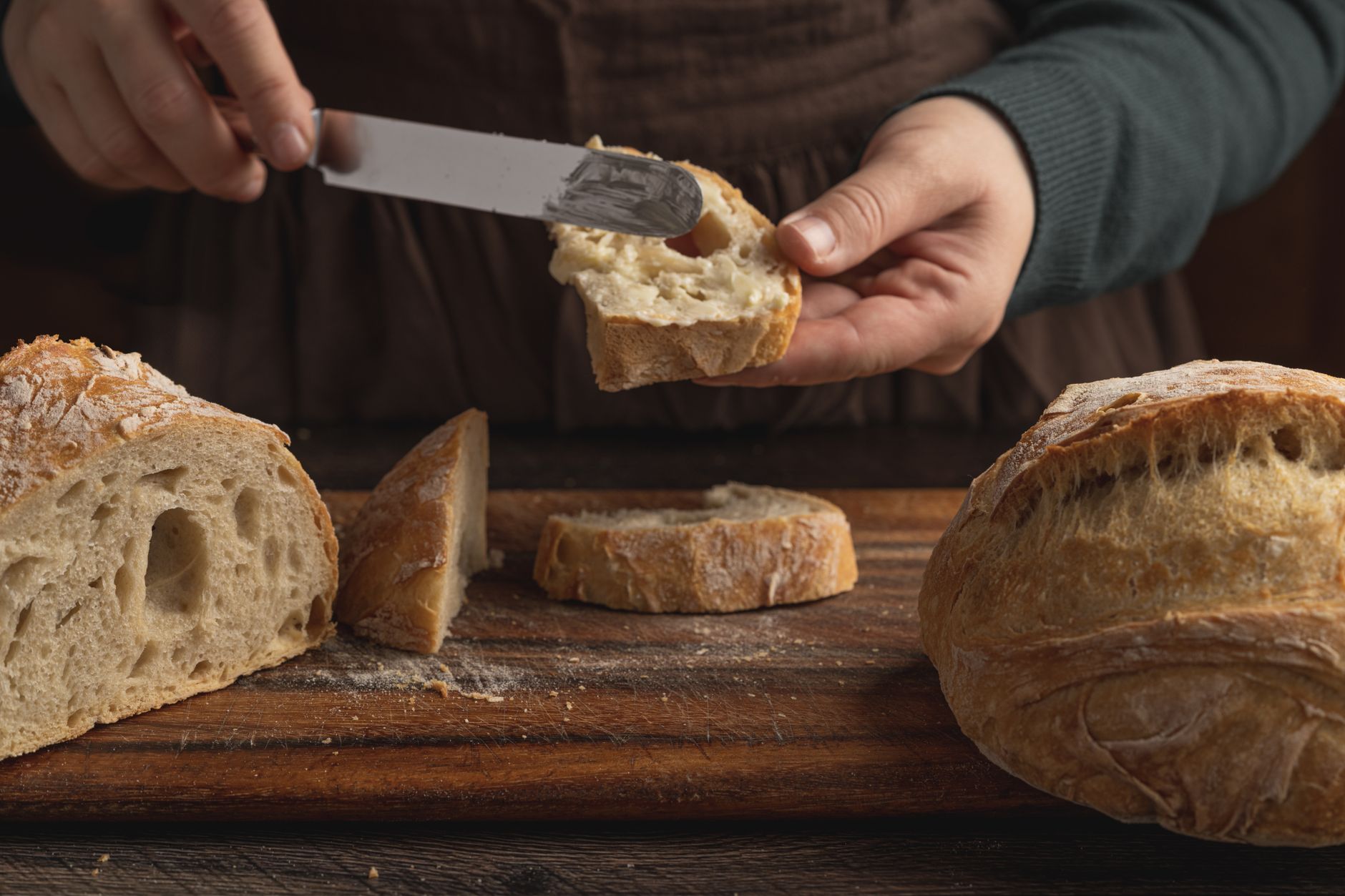Hands spreading a savoury spread on a slice of rustic bread beside a loaf on a wooden board