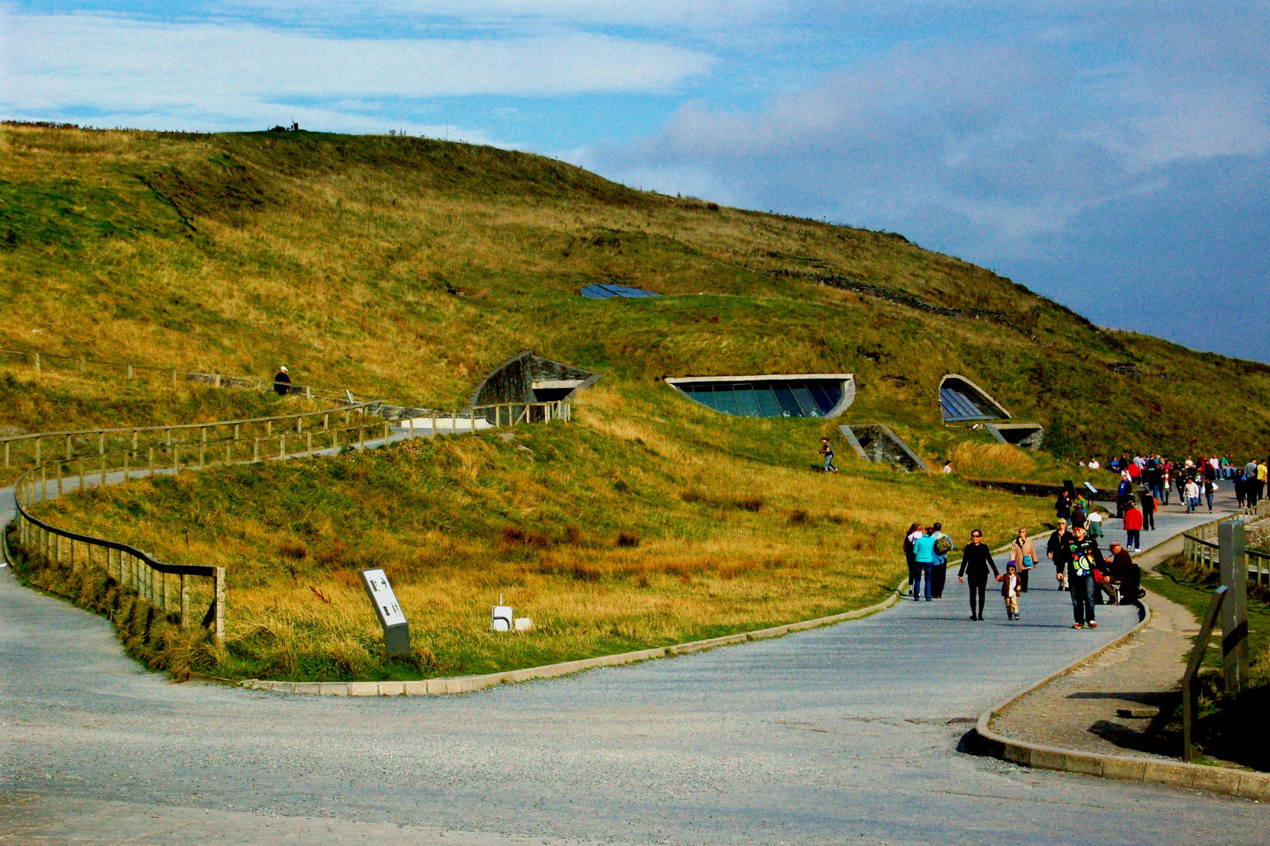 The grass-covered roof and glazed entrance of the Cliffs of Moher Visitor Centre, built into the hillside, with visitors walking along the path