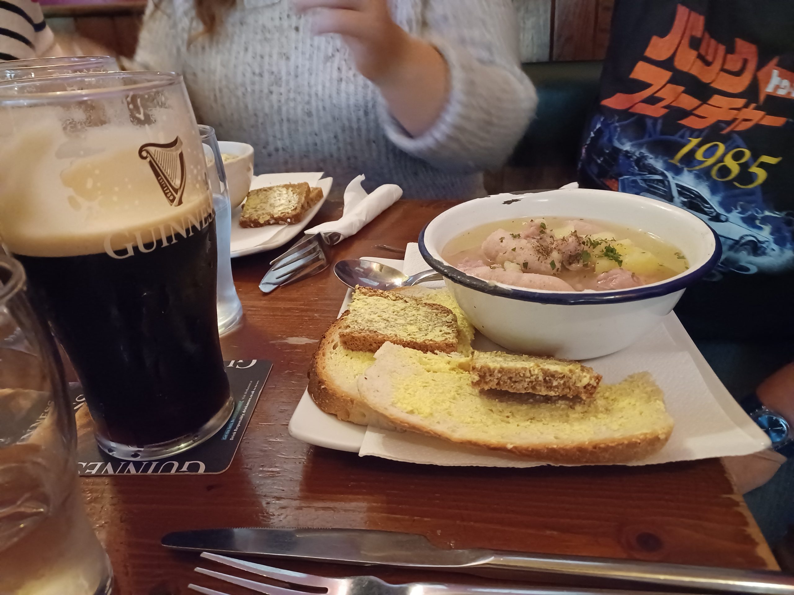 A bowl of Dublin coddle broth with bacon, sausage and potato in a clear golden broth, served with buttered soda bread and a pint of Guinness in a Dublin pub