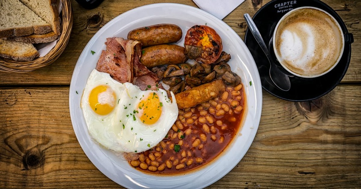 Full Irish breakfast on a white plate with fried eggs, back rashers, pork sausages, grilled tomato, mushrooms and baked beans, served with toast and coffee on a wooden table