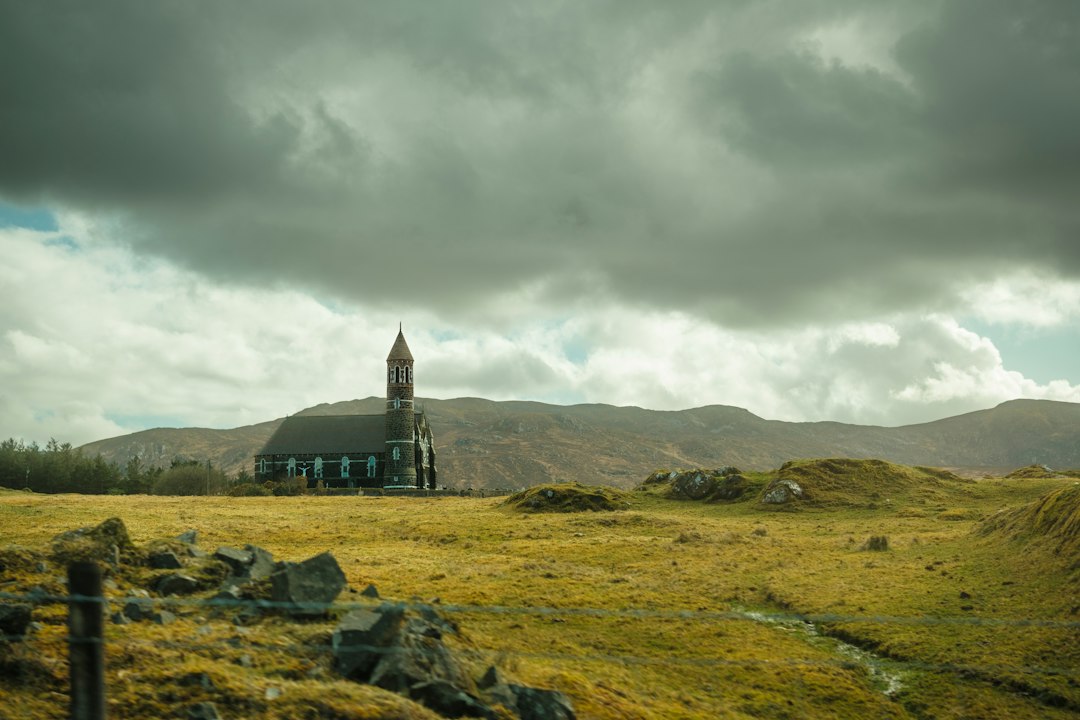 A stone church with a tall spire stands alone on green hillside pasture in Ireland under a dramatic, cloudy sky
