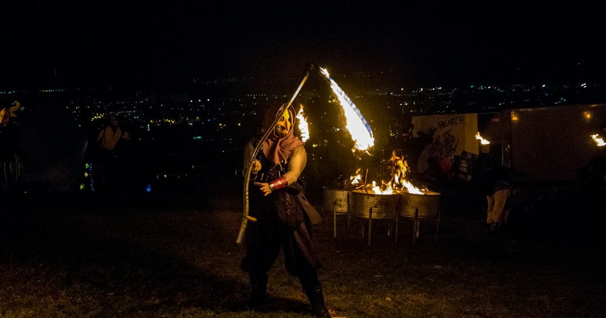 Performer swinging a flaming staff beside fire pits at a night-time outdoor event, city lights behind
