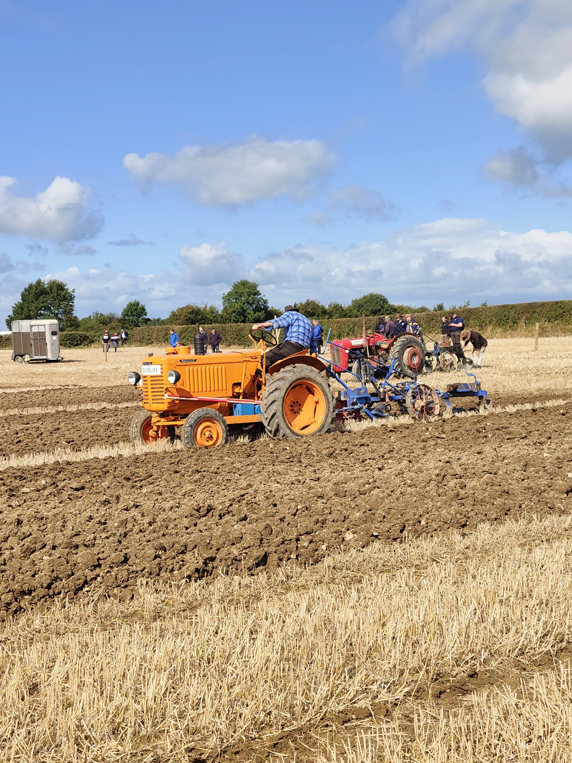 Vintage orange and red tractors pulling ploughs across a stubble field, with spectators watching at a ploughing match