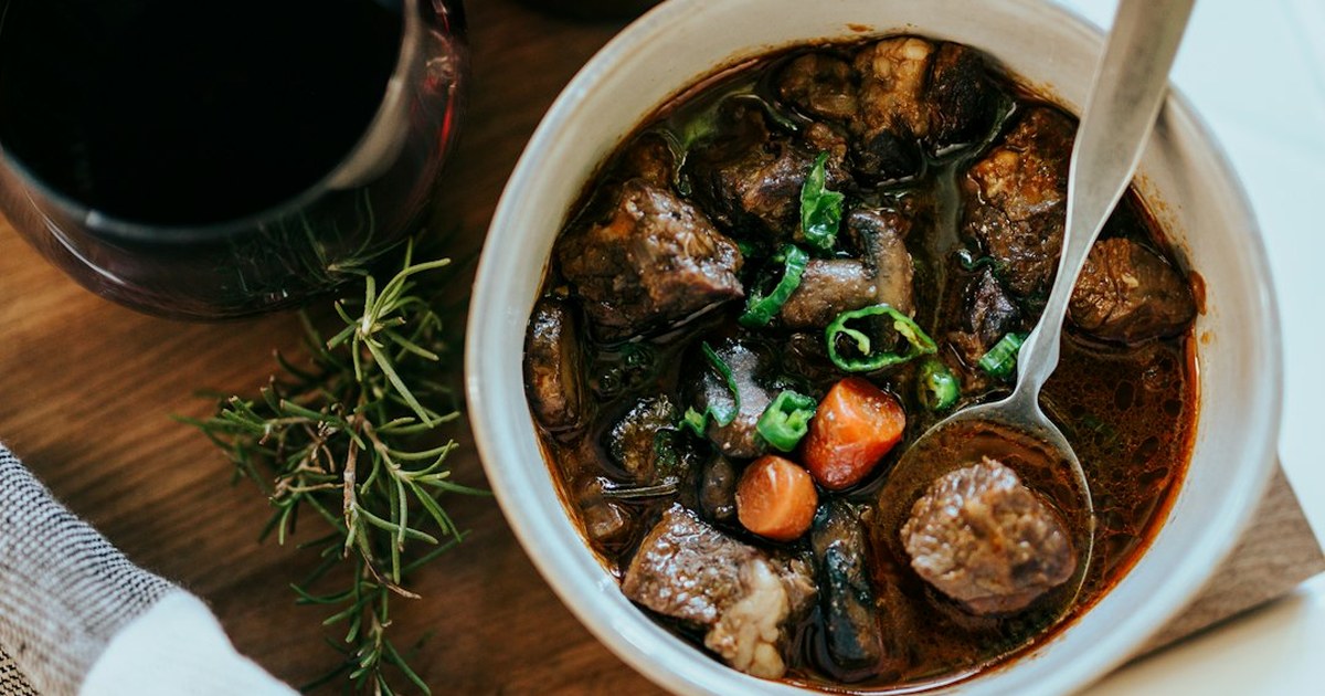 A bowl of slow-cooked oxtail stew in a rich dark stout gravy with carrot pieces and herbs, served beside a glass of stout and a sprig of fresh rosemary