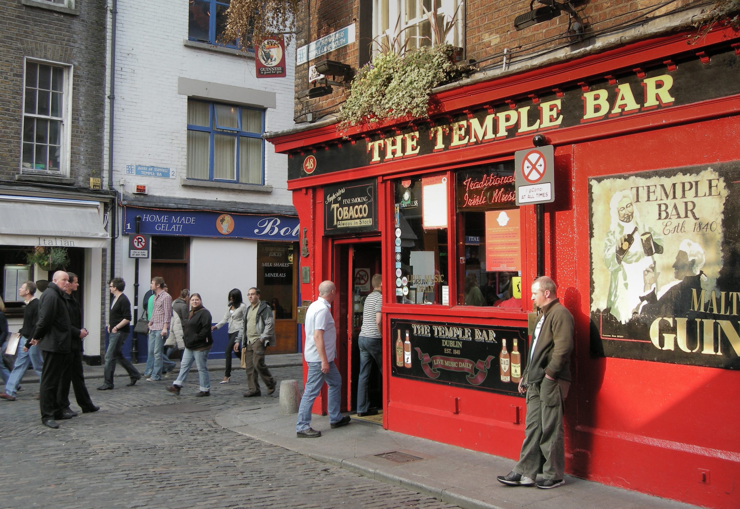 The red-fronted Temple Bar pub on a cobbled Dublin street, with a traditional Irish music sign and pedestrians passing the entrance
