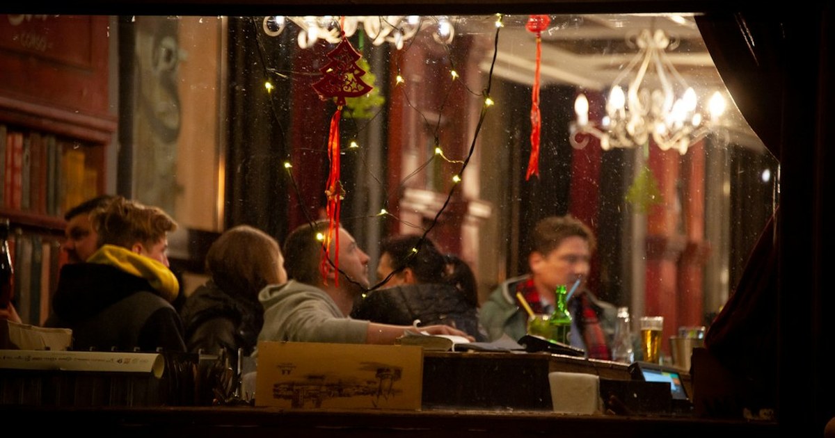 A crowded Irish pub decorated for Christmas, seen through a window, with friends gathered around the bar on Christmas Eve