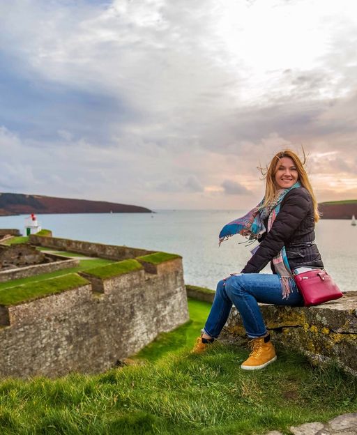 A woman sitting on ancient Irish castle ruins overlooking a harbour, with dramatic clouds above the water